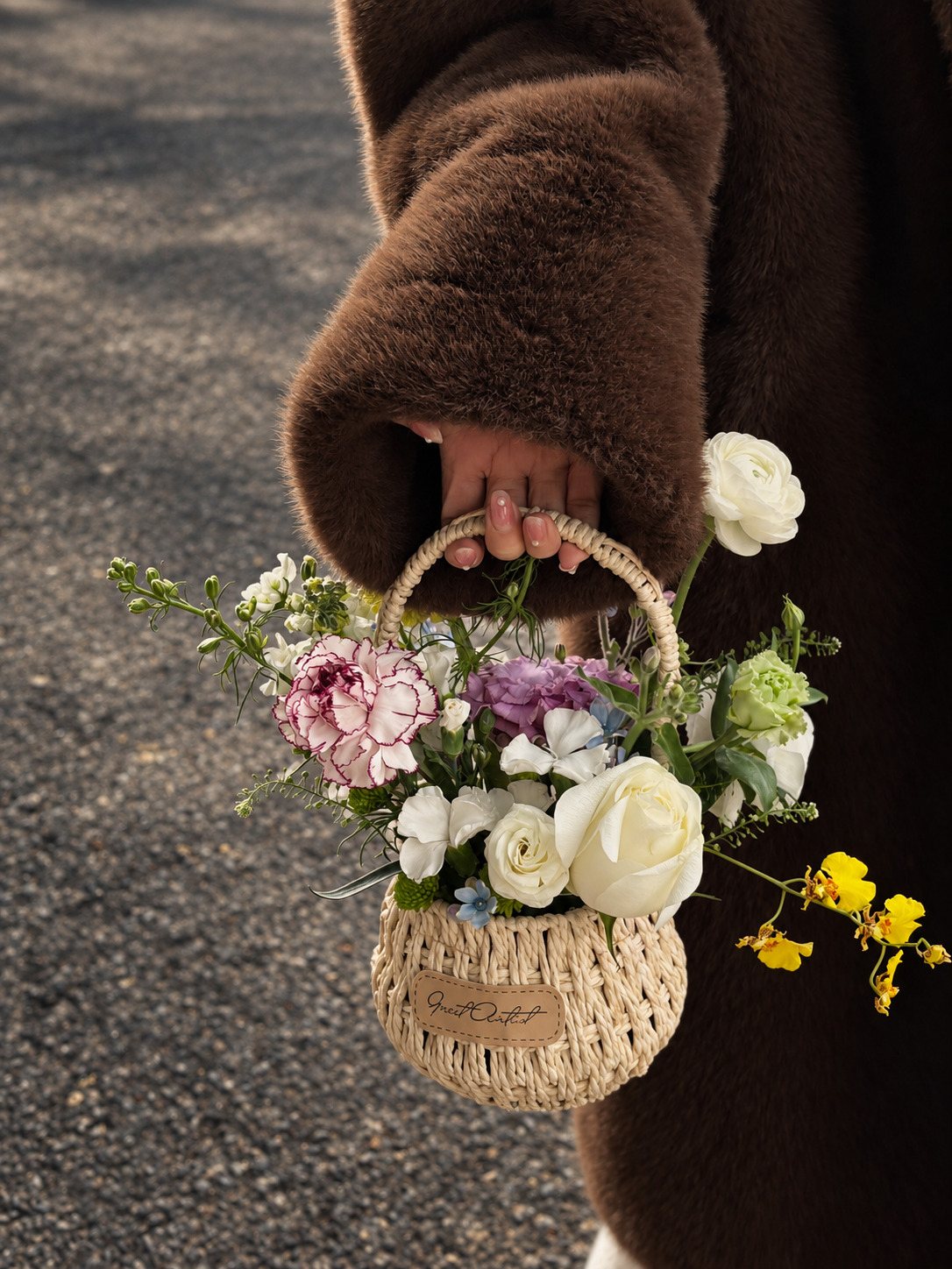 A hand in a brown faux-fur sleeve holding a small woven basket of carnations, roses, ranunculus and oncidium